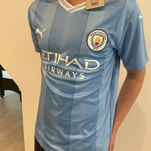 Manchester City fotbollströja 2021/22 från Puma - Snygg ljusblå Manchester City fotbollströja med vita detaljer och klubbmärke på bröstet. Puma-logga på bröstet och ärmarna, samt Etihad Airways som sponsor. Tröjan har V-ringning och är tillverkad i lätt och ventilerande material.