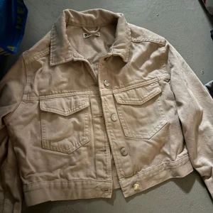Beige croppad jeansjacka från gina tricot - Säljer en beige croppad jeansjacka från gina tricot. Jackan har klassisk krage, knappar framtill och två bröstfickor med lock. Perfekt att slänga över en t-shirt eller hoodie för en cool look. Frakt från 19 kr