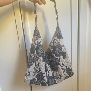 Beige y2k axelväska med blommigt mönster - Säljer en tjusig axelväska i boho-stil i beige textil med stora svarta blommor och broderade detaljer i blått. Väskan har en bred axelrem och rymlig form, perfekt för att bära med sig det viktigaste. Ger en cool och unik touch till din outfit! Väskan är varsamt använd utan defekter. Mått: 35x36 axelrem: 20cm