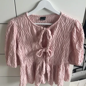 Ljusrosa blus med rosetter - Supersöt ljusrosa blus med strukturmönster och rosetter framtill. Blusen har korta puffärmar och volang nertill. Endast andvänt fåtal gånger.