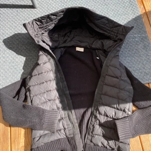 Moncler cardigan  - Snygg svart dunväst från Moncler med quiltat mönster och stickade ärmar. Västen har huva, dragkedja framtill och en extra ficka med dragkedja på ärmen. Perfekt för lager-på-lager och stilren look.