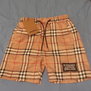 Burberry shorts med snören - Snygga beigea shorts från Burberry med klassiskt rutigt mönster i svart, vitt och rött. Shortsen har snörning i midjan och en Burberry London England-logga på ena benet. Perfekta för sommaren och har en avslappnad passform. Pris kan diskuteras i DMS.