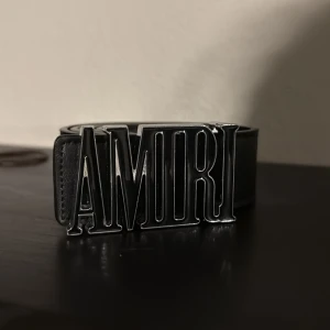 Svart bälte med stor AMIRI-logga - Säljer ett svart bälte från AMIRI med en stor, silverfärgad logotyp som spänne. Bältet är gjort i skinn och har en stilren, modern design som sticker ut med sitt tydliga varumärke framtill.