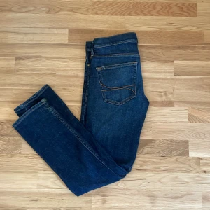 Mörkblå jeans från Hollister - Snygga mörkblå jeans från Hollister med klassisk femficksdesign och orangea sömmar. Jeansen har en skinna passform och raka ben, perfekta till vardags. Tillverkade i slitstarkt denim-material.