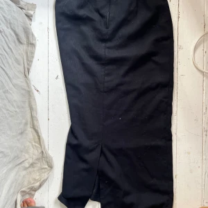 Svart långkjol med slits - Säljer en svart långkjol med slits framtill. Kjolen har hällor för bälte och dragkedja bak. Klassisk och stilren modell som passar till många olika looks.