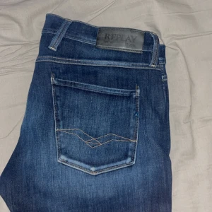 Blå Replay Anbass Jeans herr - Säljer ett par klassiska blå jeans från Replay med raka ben och snygga detaljer på bakfickorna. Jeansen har normal passform och är tillverkade i slitstarkt denim. Perfekta för dig som gillar stilrena och tidlösa jeans.