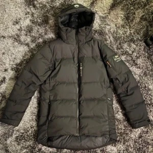 Svart dunjacka med huva från  sail recing  - Säljer en svart dunjacka från Sail recing med huva och dragkedja framtill. Jackan har lång ärm, diskret logga på bröstet och en ficka på ärmen. Perfekt för kyliga dagar. Vinter är slut men det kommer alltid till baks så köp nu för billigt 