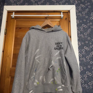 Paint splatter hoodie från DÉPT de la GALERIE - Säljer en grå hoodie från DÉPT de la GALERIE med tryck på bröstet och färgstänk i vitt, blått och gult framtill. Tröjan har huva och långa ärmar, perfekt för en avslappnad streetwear-look.
