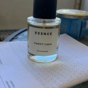 ESSNCE Sweet Cara Eau de Parfum - Fräsch parfym från ESSNCE med namnet Sweet Cara. Flaskan är genomskinlig med en enkel vit etikett och svart lock. Parfymen är en Eau de Parfum och har en stilren, modern design som passar perfekt på sminkbordet.