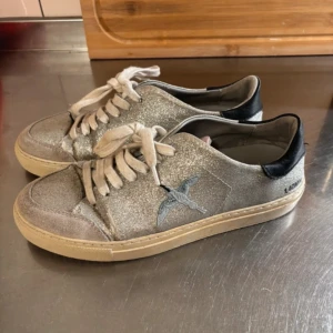 Glittriga arigatos - Säljer ett par silver glittriga Axel arigatos broderad fågel. Skorna har vita snören, beige sula och svart detalj vid hälen. Kartong och dustbag medföljer, tar även byten. Box och påse ingår 