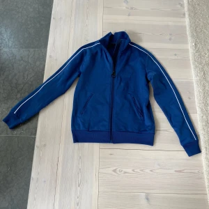 Blå track jacket Peak Performance XS - Snygg blå träningsjacka från Peak Performance i storlek XS. Jackan har vita ränder längs ärmarna, ribbade muddar och dragkedja framtill. Perfekt för en sportig look.