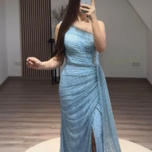 Ljusblå glittrig långklänning - Säljer en ljusblå långklänning med glittrigt tyg och oneshoulder-design. Har lagt till en extra tygbit vid axeln men går enkelt att sprätta upp! Klänningen har draperad effekt, markerad midja och en snygg slits framtill. Perfekt för bal eller fest och ger en riktigt elegant look. 