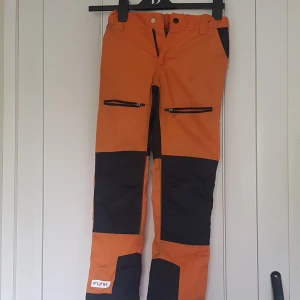 Orange täckbyxor från 157 Funk - Orange och svarta täckbyxor från 157 Funk med dragkedjor framtill och förstärkta partier på knän och bak. Perfekta för vinterns alla äventyr. Snygg kontrast mellan färgerna och praktiska fickor med dragkedja.