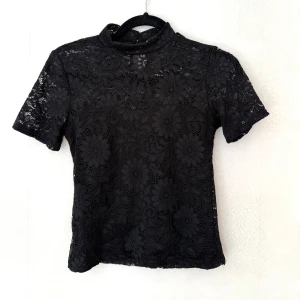 Svart spetsblus - Supersnygg svart blus från Hailys i spets med blommigt mönster. Hög hals med knapp i nacken och korta ärmar. Perfekt när du vill ha en lite extra detalj på outfiten. 