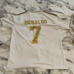 Real Madrid Ronaldo 7 matchställ - Säljer ett komplett Real Madrid matchställ med Ronaldo 7. Tröjan är vit med guldiga detaljer, klubbmärke och Fly Emirates-tryck framtill. Korta ärmar och klassiska Adidas-ränder på axlarna. Matchande shorts med klubbmärke och nummer 7 i guld. Materialet är lätt och andas. Allt ingår, pris kan diskuteras 