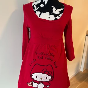 Röd Hello Kitty klänning S - Supersöt röd kortklänning med Hello Kitty Little Red Riding Hood-tryck framtill. Klänningen har långa ärmar, rund halsringning och är gjord i mjuk bomull. Perfekt för dig som älskar kawaii och vill sticka ut med en unik look.