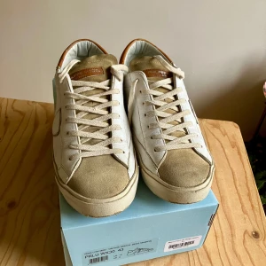 Philippe Model vita beige bruna stl 43 Originalkartong och kvitto - Snygga Philippe Model sneakers i vitt skinn med beige mockadetaljer på tå och plös. Skorna har bruna skinnpartier vid hälen och klassisk snörning. Perfekt för dig som gillar stilrena och trendiga sneakers med exklusiv känsla.Inköpta på merchsweden i november 2024, använda ca 10 gånger, nu för små. Originalkartong och kvitto medföljer.