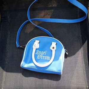 Blå axelväska Angel Dream - Säljer en blå axelväska från Angel Dream med vit text och detaljer. Väskan har bågformad design, vita handtag och lång justerbar axelrem. Materialet är syntet och den stängs med dragkedja. Perfekt för dig som vill ha en söt och unik accessoar.