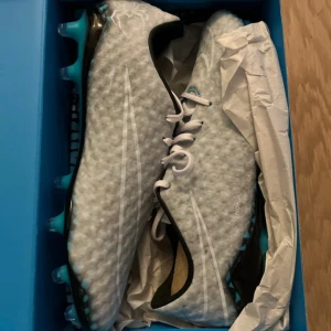 Nike Hypervenom Phantom fotbollsskor - Nike Hypervenom Phantom RGN SE FG i vitt och svart med blå detaljer. Skorna har snörning och en texturerad ovandel för bättre bollkontroll. Snygg design med blå dobbar och sportig look, perfekta för dig som vill sticka ut på planen.