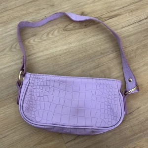 Ljuslila axelväska från ASOS Design - Säljer en ljuslila axelväska från ASOS Design med croco-mönstrad yta och guldfärgade metalldetaljer. Väskan är tillverkad i syntetmaterial och polyester, har en smidig dragkedja upptill och en bred axelrem. Perfekt för dig som vill ha en trendig accessoar med cool struktur.