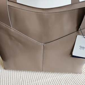 Beige Toteväska Menda Tiger of Sweden - "Oönskad present! Ursprungligen 9999 SEK på Tiger of Swedens hemsida (juni 2025), nu 4999 SEK. Rymlig Menda-väska, perfekt för jobbet, rymmer laptop och nödvändigheter. Tillverkad av högkvalitativt polido-läder, med magnetficka, säker metallblixtlås, bekväma handtag, premiumfoder utan läder och märkesficka. Stadig, stilren och funktionell i klassiska/säsongsbetonade färger."