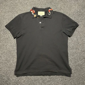 Svart Gucci kingsnake piké - Svart pikétröja från Gucci med broderad orm och bi på kragen. Klassisk pikémodell med kort ärm och knappar framtill. Materialet är bomull och kragen har snygga röda och vita detaljer som sticker ut.