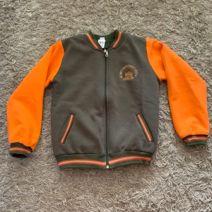 Grå och orange bomberjacka - Snygg bomberjacka i grått med orange ärmar och coola randiga detaljer på muddar och krage. Jackan har dragkedja framtill, två fickor och broderad logga på bröstet. Perfekt för dig som vill sticka ut med färg och sportig stil.