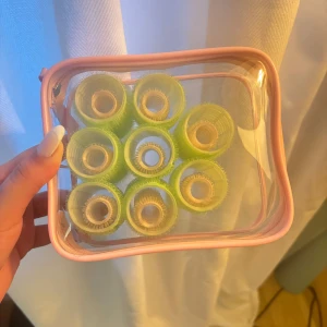 Gröna hårspolar i rosa necessär - Säljer ett set med åtta gröna hårspolar i en genomskinlig necessär med rosa detaljer och dragkedja. Perfekt för att fixa volym och lockar hemma. Necessären har Sol de Janeiro-logga på sidan och är smidig att ta med på resan.