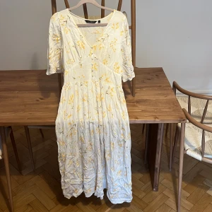 Vit blommig klänning från KappAhl - Superfin vit klänning från KappAhl med gula blommor, v-ringning och knappar framtill. Halvlånga ärmar med lätt puff och smock i ryggen. Lätt och luftig modell i bomull, perfekt för varma dagar.