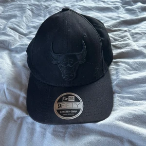 Svart Chicago Bulls keps New Era 9FIFTY - Svart keps från New Era, modell 9FIFTY Stretch-Snap, med broderad Chicago Bulls-logga framtill i svart ton-i-ton. Klassisk böjd skärm och justerbar passform. Perfekt för dig som gillar sportig streetstyle.
