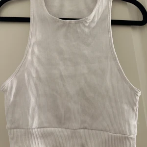 Vit ribbad linnetopp - Säljer en fräsch vit ribbad linnetopp med hög halsringning och breda axelband. Toppen är croppad och har en tight passform som sitter snyggt på kroppen. 