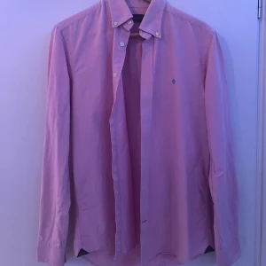 Ljusrosa skjorta Morris - Säljer en ljusrosa skjorta från Morris med klassisk krage och knappar framtill. Skjortan har lång ärm och en snygg turkos broderad lilja på bröstet. Perfekt för dig som gillar stilrena plagg med en twist. Materialet känns mjukt och skönt mot huden.