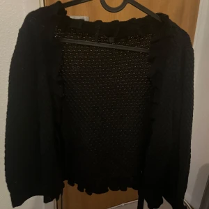 Svart stickad kofta med volangkant - Mysig svart stickad kofta med volangdetaljer längs kanterna och knytning framtill. 