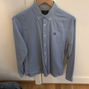 Blå skjorta från Jean Paul i bomull - Snygg blå skjorta från Jean Paul i bomull med klassisk krage och knappar framtill. Regular fit med lång ärm och diskret broderad logga på bröstet. Perfekt för dig som gillar stilrena och enkla plagg med en clean look.