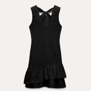 Svart kort klänning med volang och rosett - Supersöt svart kort klänning med smockad överdel, v-ringning och breda axelband. Klänningen har en rosett i ryggen och volangkant nertill. Helt oanvänd med lappen kvar