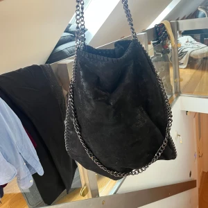 Svart axelväska med kedjedetaljer - Säljer en svart axelväska i mocka-look med grova silverfärgade kedjor som axelrem och runt kanterna. Väskan är rymlig och har en cool, slouchy form som ger en edgy vibe till din outfit.