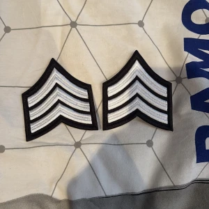 Svarta och vita sergeant tygmärken - Två stycken tygmärken i form av sergeantstreck, broderade i svart och vitt. Märkena har tydliga ränder och är perfekta att sy eller stryka fast på jackor, väskor eller hoodies för en cool militärinspirerad look.