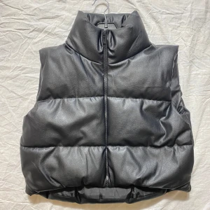 Svart dunväst i skinnimitation - Snygg svart dunväst med hög krage och dragkedja framtill. Västen har en quiltad look och är gjord i skinnimitation som ger en cool och modern vibe. Perfekt att slänga över en hoodie eller tröja för extra edge.