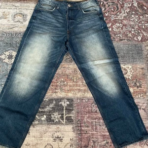 Blå loose fit jeans från H&M - Snygga blå jeans från H&M i loose fit-modell med raka ben och justerbar midja. Jeansen har klassisk femficksdesign, slitningar och ljusare partier framtill för en cool, avslappnad look. Perfekta för dig som gillar en bekväm och trendig stil.