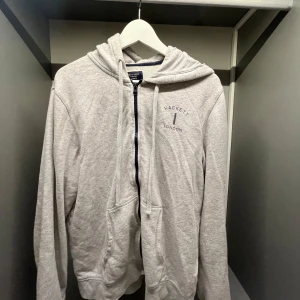 Grå hoodie från Hackett London - Snygg ljusgrå hoodie från Hackett London, modell Mr Classic. Tröjan har dragkedja, huva med snörning och två fickor framtill. Broderad logga på bröstet. Perfekt för en chill och stilren look.