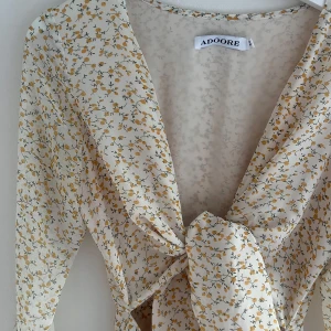 Blommig omlottklänning från Adoore XS - Supersöt kort omlottklänning från Adoore i ljusbeige/vit med små gula blommor. Klänningen har långa trumpetärmar, knyt framtill och v-ringning. Perfekt för sommarens alla tillfällen och speciellt midsommar.