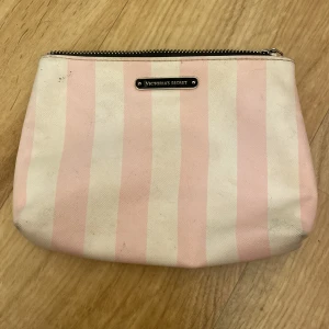 Randig necessär från Victoria's Secret - Söt necessär från Victoria's Secret i klassiskt rosa och vitt randigt tyg. Väskan har en silverfärgad dragkedja och en metallbricka med loggan framtill. Perfekt storlek för smink eller småprylar, med svartmönstrat innerfoder. Den har lite sminkfläckar inuti men inget man tänker på:)