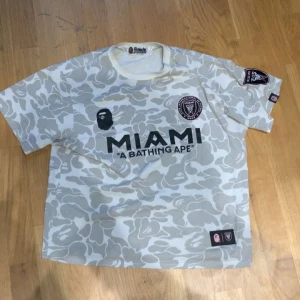 BAPE x Inter Miami camo fotbollströja - Unik Inter Miami fotbollströja från BAPE i vit och grå camouflage. Kortärmad modell med stora tryck fram och bak, inklusive MIAMI-logga, BAPE-apa och siffran 93. Patch på ärmen och detaljerad etikett. Tillverkad i bomull för skön känsla. Strl L men sitter som S-M