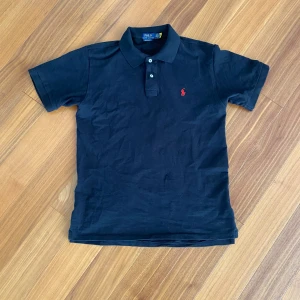 Svart Ralph Lauren Piké - 👕 Märke: Polo Ralph Lauren 📏 Storlek: M (Slim Fit) 🎨 Färg: Svart med röd logga ✨ Skick: Helt ny – aldrig använd  Klassisk svart pikétröja från Polo Ralph Lauren med den ikoniska röda loggan. Aldrig använd – endast provad. Snygg, tidlös passform i mjuk bomull.