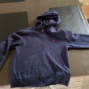 Mörkblå hoodie från Polo Ralph Lauren junior  - Snygg mörkblå hoodie från Polo Ralph Lauren med dragkedja och klassisk röd logga på bröstet. Tröjan har huva, känguruficka och långa ärmar. Perfekt för en chill och stilren look.