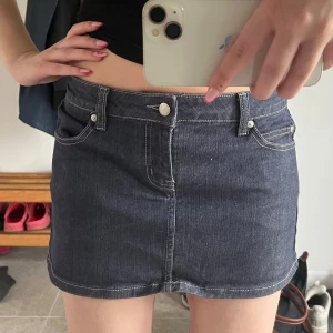 Lågmidjad jeanskjol med fickor - Säljer en klassisk mörkblå jeanskjol med fickor och tydliga kontrastsömmar. Kjolen är kort och har bälteshällor samt knapp och dragkedja framtill. Perfekt för dig som gillar en enkel och tidlös denimlook.