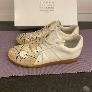 Maison Margiela sneakers med färgstänk - Unika vita sneakers från Maison Margiela med handmålade färgstänk i gult, rött, svart, grönt och rosa. Skorna har klassisk låg siluett, snörning och gummisula. Tillverkade i skinn och har en konstnärlig vibe som sticker ut i mängden. Jag tvättar dem innan frakt och skickar även bild på när de är tvättade. Priset ej satt i sten!
