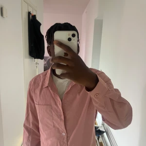 Rosa skjorta med knappar - Säljer en stilren rosa skjorta med klassisk krage och bröstficka. Skjortan har långa ärmar och knäppning framtill. Perfekt för dig som vill ha en enkel men snygg look. Materialet känns lätt och passar bra till många outfits.