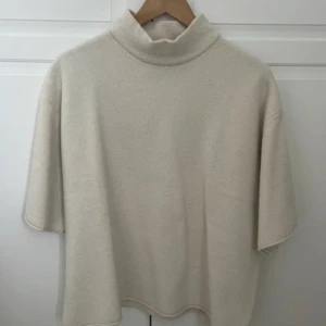 Beige polotröja med kort ärm - Beige polotröja med lös passform och korta ärmar. Tröjan har en hög krage och är tillverkad i ett mjukt, stickat material som känns riktigt skönt mot huden. Perfekt för lager-på-lager och enkel att styla till olika outfits.