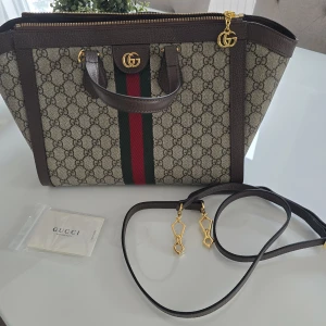 Gucci GG Supreme axelväska med rem - Säljer en lyxig Gucci axelväska i GG Supreme-mönster med bruna skinn-detaljer, guldiga metalldetaljer och klassiska gröna/röda ränder. Väskan har både handtag och avtagbar axelrem. Perfekt för dig som vill ha en ikonisk och trendig väska med plats för allt du behöver. Priset kan diskuteras vid snabb affär. 